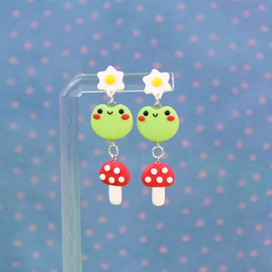 May include: A pair of whimsical dangle earrings featuring a green frog design with red cheeks, a red mushroom, and a white daisy. The earrings are suspended from a silver-colored chain. The background is a light blue with a polka dot pattern.