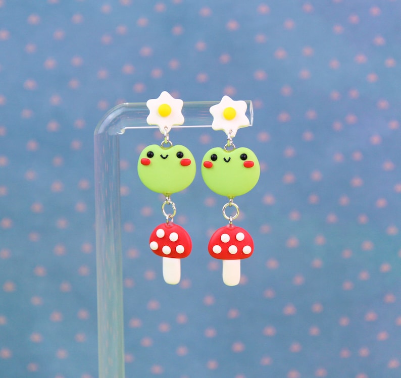 May include: A pair of whimsical dangle earrings featuring a green frog design with red cheeks, a red mushroom, and a white daisy. The earrings are suspended from a silver-colored chain. The background is a light blue with a polka dot pattern.