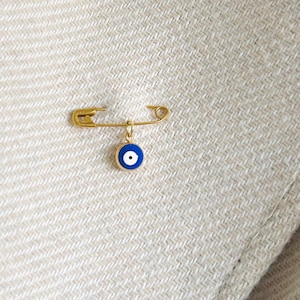 May include: A gold safety pin with a blue and white evil eye charm attached. The pin is on a beige fabric background.