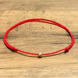 May include: A red string bracelet with a small gold bead in the centre. The bracelet is knotted at each end. The jewellery is displayed on a wooden surface.