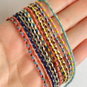 May include: A collection of colorful braided cords with gold thread accents. The cords are arranged in a stack and are approximately 1/4 inch wide.