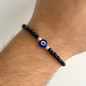 May include: A black beaded bracelet with a blue evil eye charm. The charm has a white centre and silver accents.