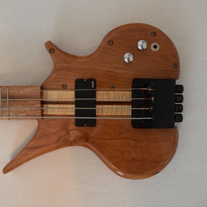 May include: A wooden electric bass guitar with a unique body shape and a black pickup. The guitar has a natural wood finish and a black bridge.