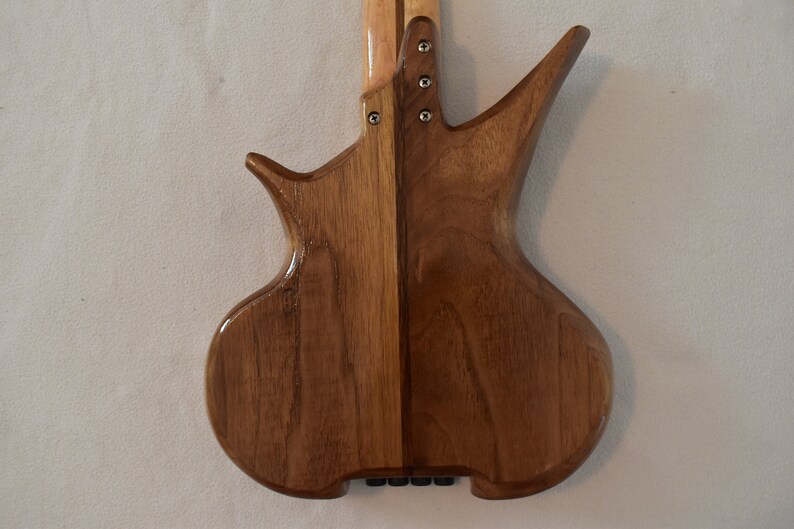 May include: A wooden electric bass guitar with a unique body shape and a light brown finish. The body is made of two pieces of wood, with a dark brown stripe down the center.