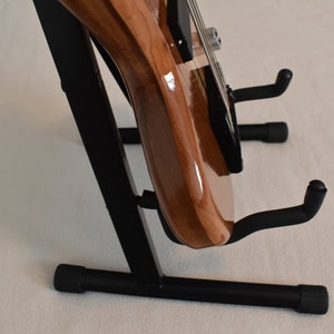 May include: A brown electric bass guitar with a black pickguard is displayed on a black metal guitar stand. The stand has a triangular base with three legs and two arms that hold the guitar in place.