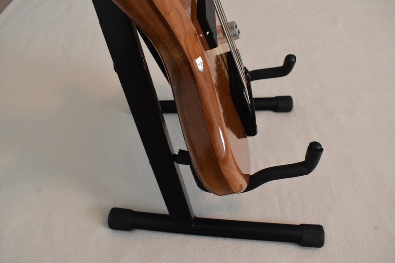 May include: A brown electric bass guitar with a black pickguard is displayed on a black metal guitar stand. The stand has a triangular base with three legs and two arms that hold the guitar in place.