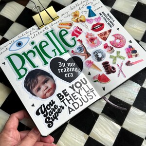The image is a hand holding a scrapbook page with various stickers and a photo of a child's face. The page has a black and white checkered background, and the stickers include a birthday cake, a hot dog, a donut, and other colorful and playful elements. The phrase "You are Super Adjust" is also visible on the page.