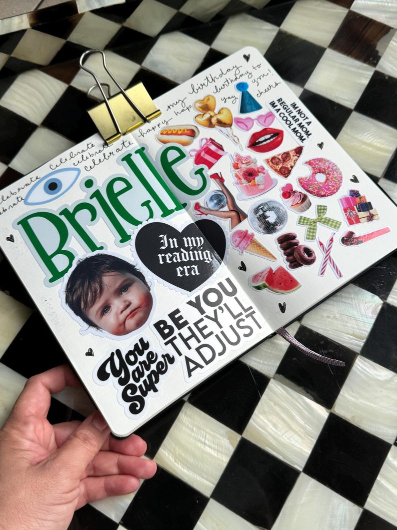 The image is a hand holding a scrapbook page with various stickers and a photo of a child's face. The page has a black and white checkered background, and the stickers include a birthday cake, a hot dog, a donut, and other colorful and playful elements. The phrase "You are Super Adjust" is also visible on the page.