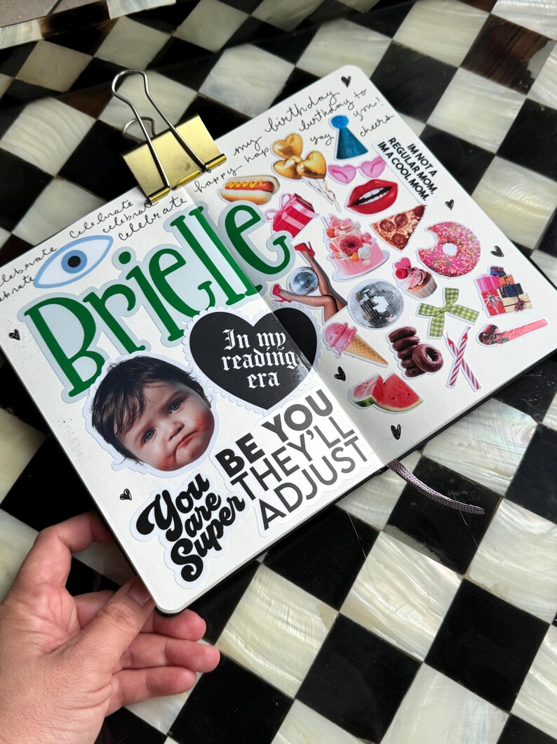 The image is a hand holding a scrapbook page with various stickers and a photo of a child's face. The page has a black and white checkered background, and the stickers include a birthday cake, a hot dog, a donut, and other colorful and playful elements. The phrase "You are Super Adjust" is also visible on the page.