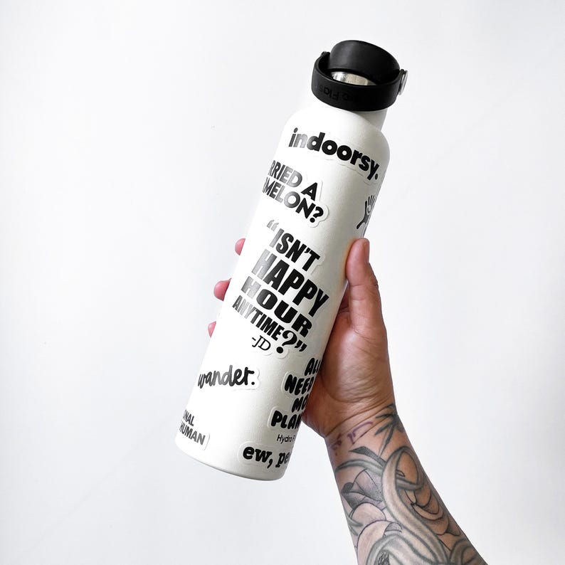 A person's hand holding a white water bottle with various text and stickers on it.