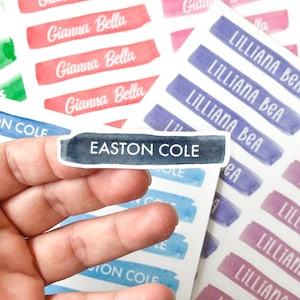 May include: A collection of watercolor-style name labels in various colors. One label, held by a hand, reads "EASTON COLE" in white text on a dark blue background. Other labels display names like "Gianna Bella" and "Lilliana Bea".