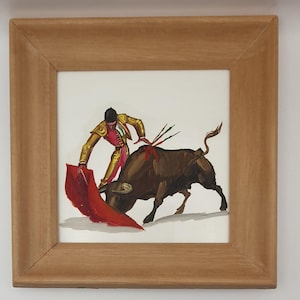 May include: Framed artwork depicting a bullfight scene. A matador in a gold and pink suit faces a brown bull with a red cape. The artwork is in a light brown wooden frame.