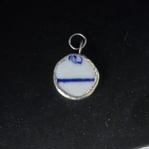 May include: A silver pendant with a round, white porcelain charm featuring a blue and white design. The charm is framed with a silver border.