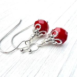 May include: A pair of drop earrings featuring faceted, round, bright red beads. Each bead is topped with a decorative, scalloped silver-tone metal cap and connected to a simple silver-tone wire hook by a coiled silver-tone metal wire stem.