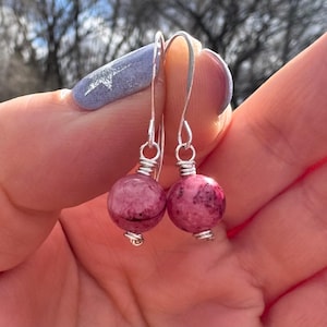 May include: A pair of dangle earrings featuring round, pink gemstone beads with silver-colored hooks. The beads have a speckled appearance, and the hooks are simple, curved wires. The earrings are held up against a blurred background.