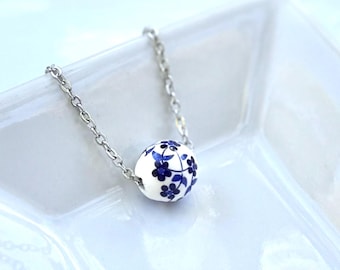 May include: A delicate necklace featuring a white ceramic bead with a blue floral pattern. The bead is spherical and suspended on a silver chain. The necklace is displayed on a white, square dish.