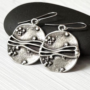 May include: A pair of circular silver-toned earrings with a textured, oxidized finish. Each earring features a prominent horizontal wavy line across the center and small raised spherical accents scattered across the surface, suspended by hooks.