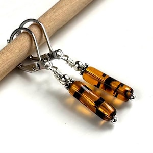May include: A pair of dangle earrings featuring cylindrical amber-colored glass beads with black horizontal stripes. The beads are accented with small silver-toned metal spacers and beads, suspended from silver-toned metal leverback ear wires.