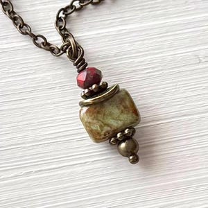 May include: A necklace featuring a square, mottled green and brown stone bead pendant accented with a small, faceted red bead and antique brass-toned metal spacers. The pendant hangs from a matching antique brass-toned metal link chain.