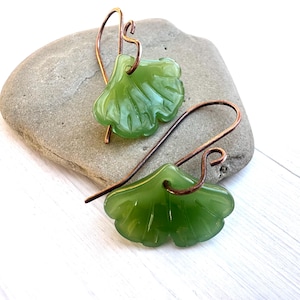May include: A pair of green leaf-shaped earrings with copper-colored ear wires. The earrings are resting on a gray stone against a white wooden background. The leaves have a glossy finish.