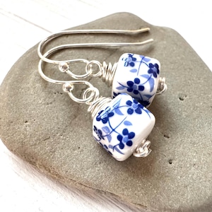 May include: A pair of dangle earrings featuring square ceramic beads with a white base and a blue floral pattern. The beads are attached to silver-toned metal ear wires with coiled wire accents, creating a simple and elegant jewelry design.