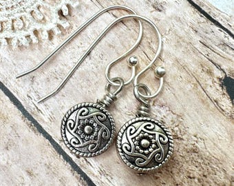 May include: A pair of silver-toned earrings with a circular design. Each earring features a detailed, embossed pattern with a central floral element and swirling accents. The earrings have a hook closure and are displayed on a wooden surface.