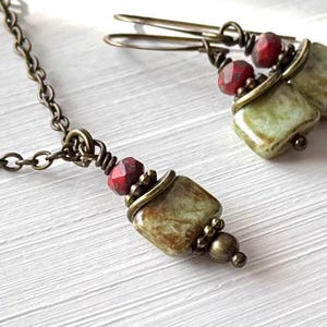May include: A jewelry set consisting of a necklace and matching earrings featuring square, mottled green beads, faceted dark red beads, and antique bronze-toned metal components. The necklace has a link chain, and the earrings have hook-style wires.