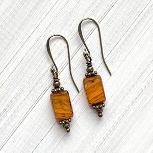 May include: A pair of handcrafted earrings featuring rectangular orange glass beads with a swirled pattern. The beads are accented with antique bronze-colored metal beads and ear wires. The earrings are displayed on a white wooden surface.