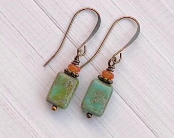 May include: A pair of handcrafted earrings featuring rectangular, turquoise-colored beads with a distressed finish. The earrings have bronze-colored ear wires and are accented with small orange beads and gold-colored metal details.