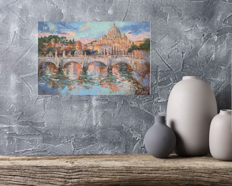 May include: An impressionistic painting of a European cityscape featuring a domed cathedral, arched bridges, and a river. The artwork uses a palette of blues, oranges, and pinks, with visible brushstrokes. The painting is displayed on a textured gray wall.