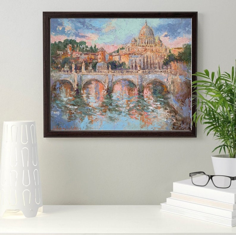 May include: An oil painting in a dark brown frame depicts a cityscape with a bridge over water. The painting features a large domed building and other structures, with reflections in the water. The color palette includes blues, pinks, and browns.