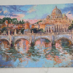 May include: An oil painting depicting a cityscape with a bridge over water. The painting features a large domed building, possibly a cathedral, and other buildings in warm tones. The sky is a mix of blue, pink, and purple.