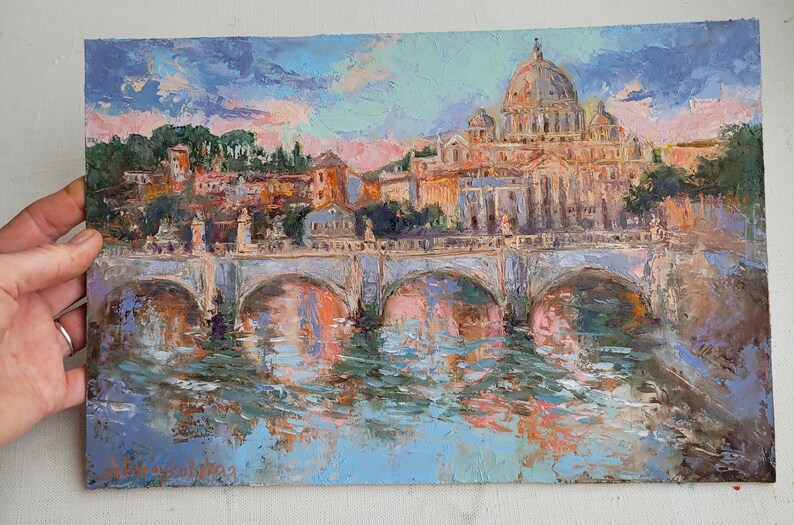 May include: An oil painting depicting a cityscape with a bridge over water. The painting features a large domed building, possibly a cathedral, and other buildings in warm tones. The sky is a mix of blue, pink, and purple.