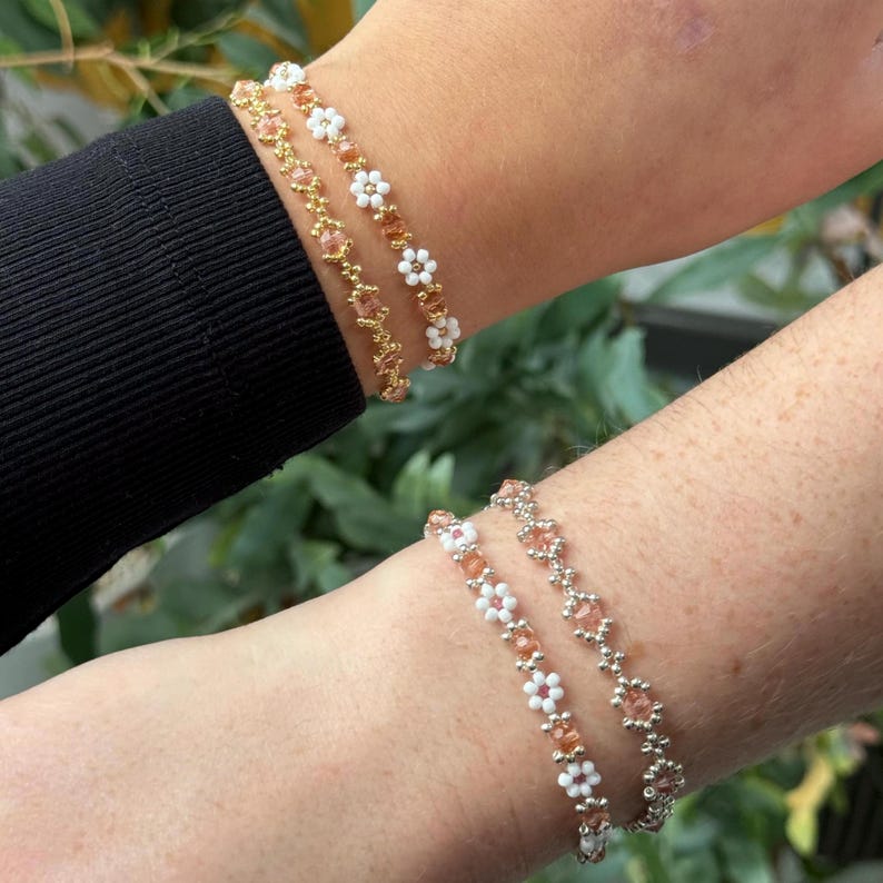 May include: Two beaded bracelets with white flower accents. The bracelets are made with small, light pink and gold beads. The bracelets are stacked on a person's wrist.