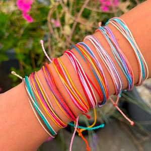 May include: A collection of multiple thin, waterproof string bracelets in a vibrant array of colors including turquoise, brown, yellow, pink, orange, red, purple, white, and light blue, layered together to create a stacked appearance on the wrist.