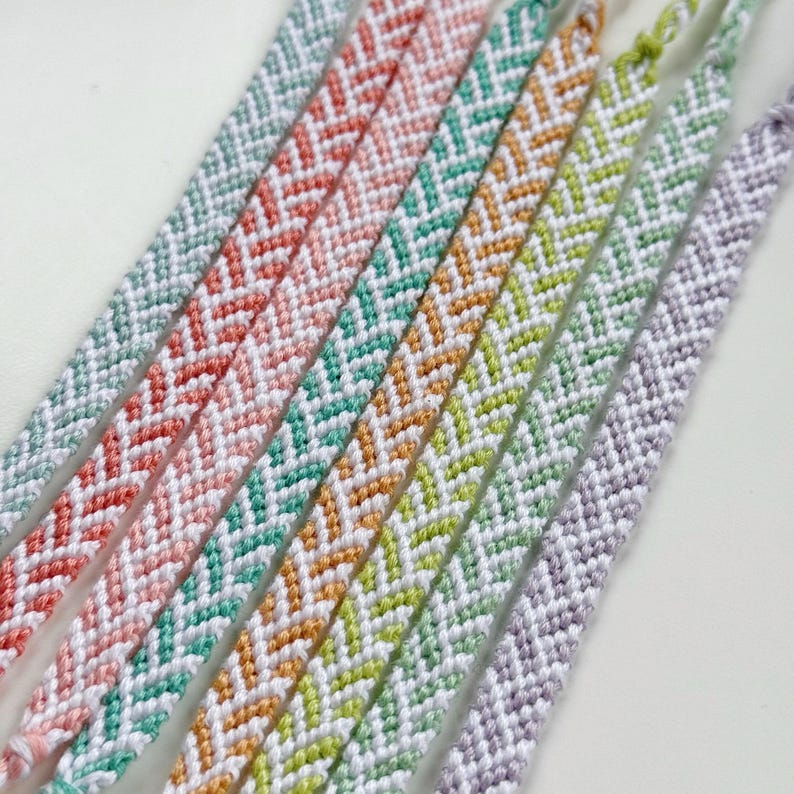 May include: Close-up of several handmade friendship bracelets in various colors, including light blue, coral, teal, yellow, and lavender. Each bracelet features a chevron pattern in white, creating a textured and colorful accessory.