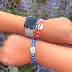 May include: Two wrists each adorned with a seashell bracelet and a smartwatch. One bracelet features a red and pink chevron pattern, the other a blue and white chevron pattern. Both bracelets have a white seashell in the center.
