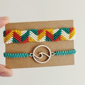 May include: Two handmade friendship bracelets. One features a chevron pattern in yellow, red, teal, and white. The other is teal with a silver wave charm. Both are displayed on a brown card.