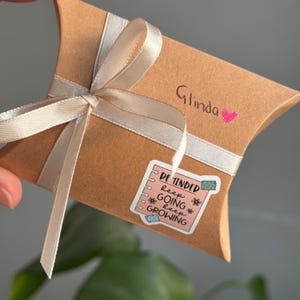 May include: A brown cardboard gift box with a white satin ribbon tied in a bow. The box has a pink sticker with the text "REMINDER keep GOING keep GROWING" and a pink heart with the name "Glinda" written on it.