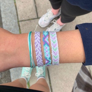 May include: A wrist adorned with several colorful friendship bracelets. The bracelets feature shades of purple, blue, and green, with intricate woven patterns. A turquoise and white string bracelet is also visible. The background shows feet wearing sneakers.