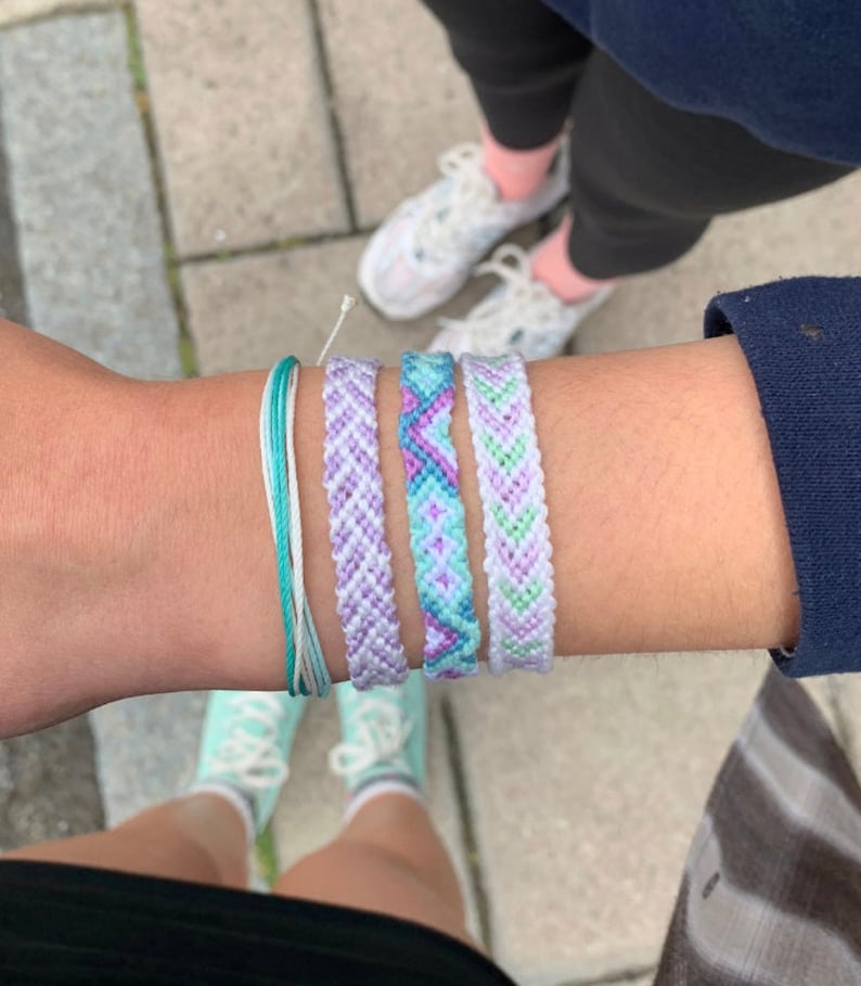 May include: A wrist adorned with several colorful friendship bracelets. The bracelets feature shades of purple, blue, and green, with intricate woven patterns. A turquoise and white string bracelet is also visible. The background shows feet wearing sneakers.