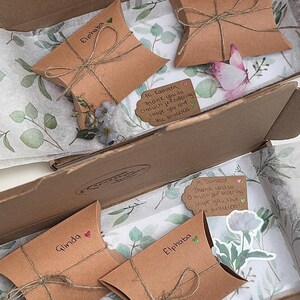 May include: A cardboard box with four brown paper gift bags inside. The bags are tied with twine and have handwritten names on them: Glinda, Elphaba, Glinda, and Elphaba. Each bag has a small decorative element, such as a flower or a butterfly. The box is lined with white tissue paper with a green floral pattern.