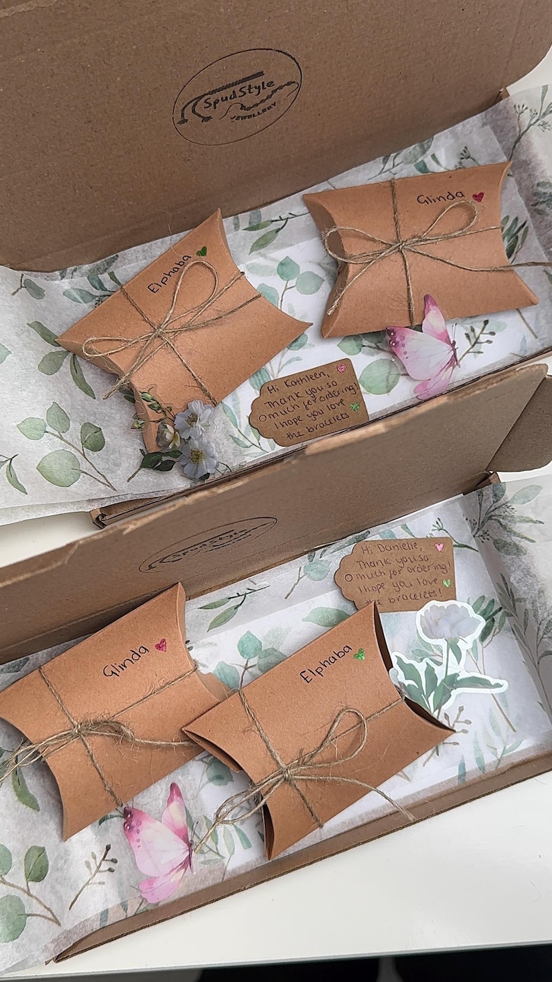 May include: A cardboard box with four brown paper gift bags inside. The bags are tied with twine and have handwritten names on them: Glinda, Elphaba, Glinda, and Elphaba. Each bag has a small decorative element, such as a flower or a butterfly. The box is lined with white tissue paper with a green floral pattern.