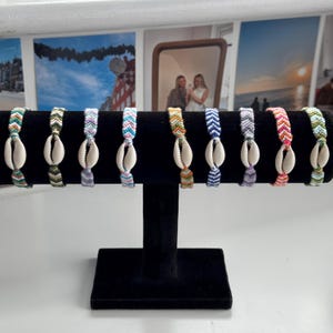 Könnte beinhalten: Zehn bunte Freundschaftsarmbänder mit weißen Muschelanhängern, die auf einem schwarzen Ständer ausgestellt sind. Die Armbänder sind aus buntem Stickgarn gefertigt und haben ein Chevron-Muster.