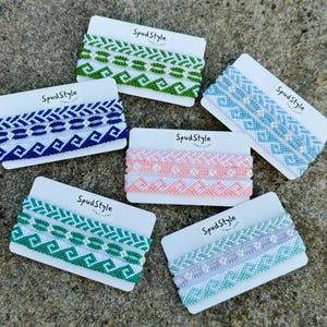 May include: A selection of woven friendship bracelets in various colours, including green, blue, pink, and purple, displayed on white cards. Each card is labelled with the text "SpudStyle". The bracelets feature detailed patterns.