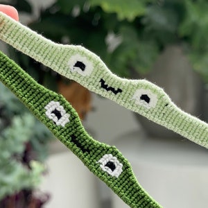 May include: Two green friendship bracelets with white and black embroidered eyes. The bracelets are woven with a simple stitch pattern.