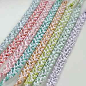 May include: A collection of handmade friendship bracelets in various colors, including pink, green, yellow, and purple. Each bracelet features a woven chevron pattern, showcasing a mix of white and vibrant hues. The bracelets are displayed on a white surface.
