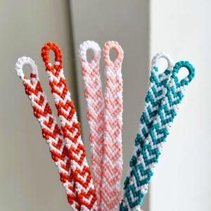 May include: Close-up of five handmade friendship bracelets in various colours. The bracelets feature a chevron pattern with white, orange, pink, and teal threads. Each bracelet has a loop at the top for tying.