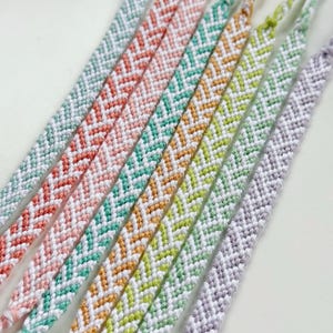 May include: Close-up of several colorful friendship bracelets. Each bracelet features a chevron pattern in various colors, including pink, teal, orange, yellow, and purple. The bracelets are made of woven thread.