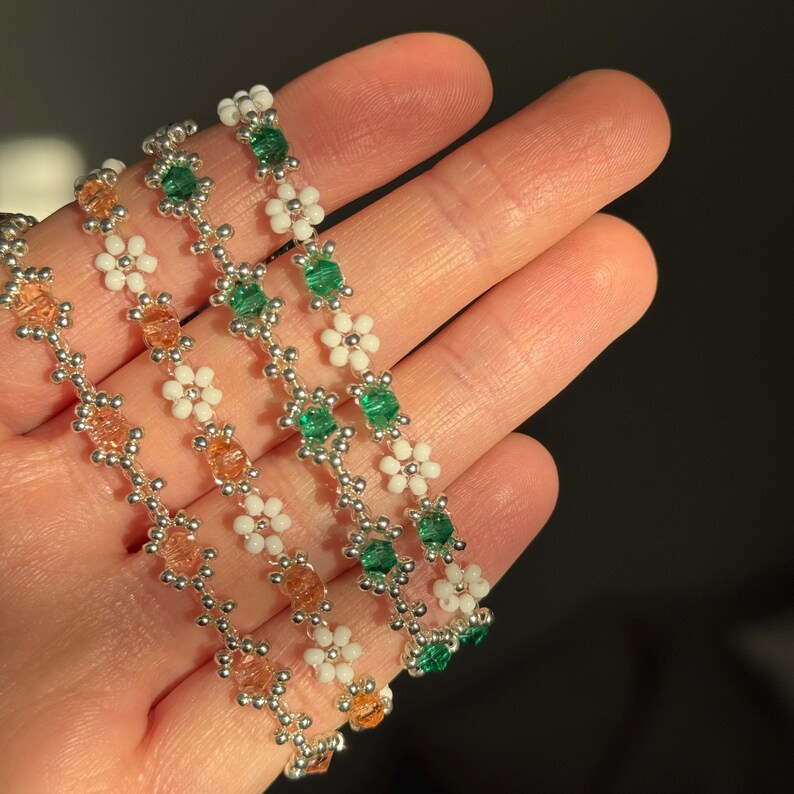 May include: Three beaded bracelets with silver beads, white flower beads, and green beads. The bracelets are stacked on top of each other.